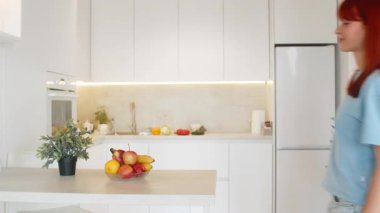 Bardan elma toplayan, meyve kasesinden elma toplayan, ısıran, gülümseyen genç bir kadın. Mutfaktaki kızıl saçlı kız taşınıyor. Mutlu adımlar. Taze meyve atıştırmalığıyla.