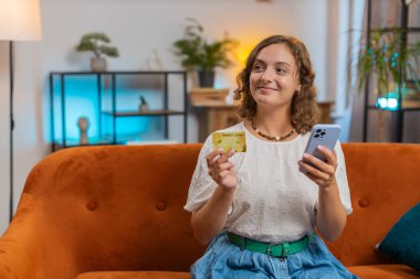 Para transfer ederken kredi kartı akıllı telefonu kullanan gülümseyen genç bir kadın, kapalı bir evde çevrimiçi alışveriş nakitsiz yemek siparişi veriyor. Kanepede oturan mutlu kız. Yaşam Tarzları