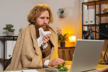 Sağlık, soğuk ve insanların kavramları. Battaniyeye sarılı hasta kızıl saçlı işadamı ofis masasındaki laptopta çalışıyor. Soğuktan titreyen sağlıksız serbest çalışan biri. Coronavirus karantinası