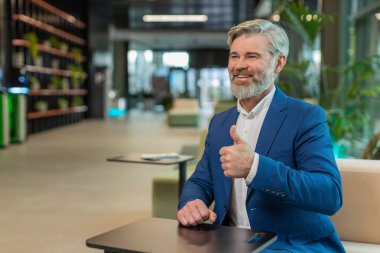 Gibi. Takım elbiseli mutlu, orta yaşlı bir işadamı kameraya bakıp olumlu bir işaret veriyor. Modern ofis lobisinde oturmuş olumlu bir izlenim veriyor. İş insanları.