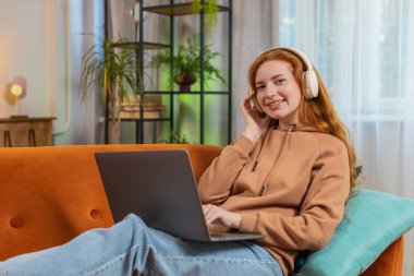 Kablosuz kulaklıklı mutlu, gevşemiş Kafkas genç kadın evinde oturuyor ve dizüstü bilgisayar ağı üzerinde çalışırken disko müziği dinliyor. Kızıl saçlı kız hafta sonu aktiviteleri.