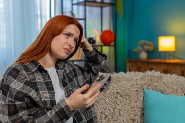 Evde elinde akıllı telefon olan genç bir kadın kötü haberden sonra üzgün bir suratla ekrana bakıyor. Kanepedeki beyaz saçlı kız üzgün hissediyor, mesajlara duygusal tepki veriyor ya da beklenmedik sonuçlar veriyor.