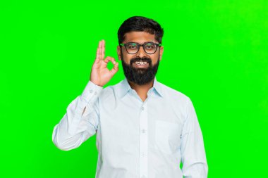 Hintli adam neşeli bir gülümsemeyle iyi el hareketi yapar. Anlaşmayı ve dostça enerjiyle olumlu tutumu ifade eder. Yeşil krom anahtar arka planında izole edilmiş Arap adam onay ve destek gösteriyor