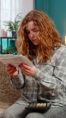 Kadın evde hesap makinesi kullanarak faturaları ve faturaları dikkatle incelerken harcamaları hesaplıyor. Kanepede mali belgeleri düzenleyen, bütçe planlayan ve harcama kontrolü yapan beyaz saçlı bir kız.