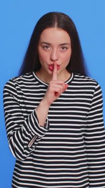 Sessiz olun lütfen. Beyaz kadın işaret parmağını dudaklarına bastırıyor. Sessiz jest işareti yapıyor. Sırları söyleme, dedikodu yapma, mahremiyetten bahsetme. Güzel genç kız mavi arka planda izole edilmiş.