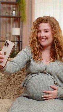 Hamile genç bir kadın akıllı telefonuyla video görüşmesi yapıyor, karnına hafifçe dokunuyor, kanepesinde mutlulukla gülümsüyor. Özel anları ve duygusal bağlantıyı paylaşan kız blogcu.