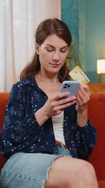 İnternetten alışveriş yapan mutlu genç bir kadın. Gülümseyen beyaz kız elinde bankacılık kredi kartı tutuyor evdeki koltukta mobil uygulama doğrulama sürecinde yer alıyor..