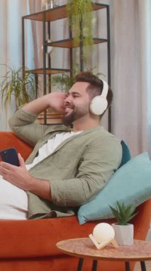 Kablosuz kulaklıklı, akıllı telefonlu mutlu genç adam evinde kanepeye uzanmış, en sevilen enerjik disko dans müziğini dinliyor. Hafta sonu eğlence aktiviteleri.