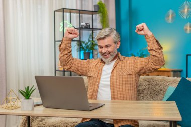 Laptop kullanan mutlu beyaz adam önemli bir e-posta mesajına ulaşıyor. İyi haber. Tatmin olmuş orta yaşlı bir adam evinde oturmuş sevinçle bağırıyor, oyunu kazandıktan sonra ellerini kaldırıyor.