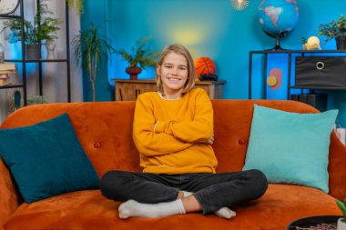 Kafkasyalı genç çocuk evde samimi bir gülümsemeyle bir portre için poz veriyor, rahatlatıcı. Kollarını kavuşturup koltukta rahatça oturur, kameraya bakar, mutluluk ve kendine güvenini gösterir.