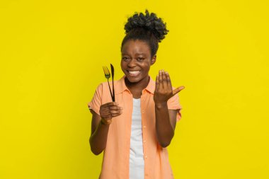Afro-Amerikalı kadın çatal ve bıçak tutuyor davetkar bir hoş geldin jesti ile, dostluk saçıyor. Sarı arka plandaki siyah kız, ortak yemek deneyiminin zevkini vurguluyor