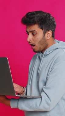 Memnun olmayan Hintli adam dizüstü bilgisayar kullanıyor. Bilgisayarda arama yapıyor. Piyango kötü sonuçlara şaşırıyor. Pembe arka planda izole edilmiş Arap adam.