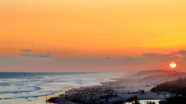 Altın turuncu gökyüzü, okyanus dalgaları ve akşamdan zevk alan insanların siluetleri ile güzel tropikal plaj günbatımı. Seyahat, turizm ve doğa için mükemmel sakin bir sahil manzarası..