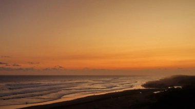 Altın turuncu gökyüzü, okyanus dalgaları ve akşamdan zevk alan insanların siluetleri ile güzel tropikal plaj günbatımı. Seyahat, turizm ve doğa için mükemmel sakin bir sahil manzarası..