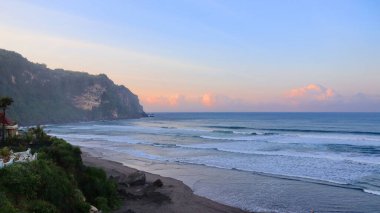 Tropikal plajın çarpıcı deniz manzarası kıyıya vuran dalgalar, dramatik sahil kayalıkları ve günbatımında pastel gökyüzü. Seyahat, doğa ve manzara için mükemmel..