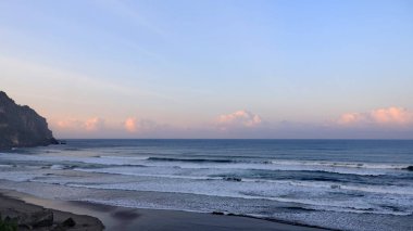 Tropikal plajın çarpıcı deniz manzarası kıyıya vuran dalgalar, dramatik sahil kayalıkları ve günbatımında pastel gökyüzü. Seyahat, doğa ve manzara için mükemmel..