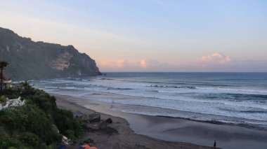 Tropikal plajın çarpıcı deniz manzarası kıyıya vuran dalgalar, dramatik sahil kayalıkları ve günbatımında pastel gökyüzü. Seyahat, doğa ve manzara için mükemmel..