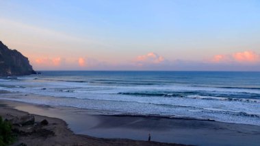 Tropikal plajın çarpıcı deniz manzarası kıyıya vuran dalgalar, dramatik sahil kayalıkları ve günbatımında pastel gökyüzü. Seyahat, doğa ve manzara için mükemmel..