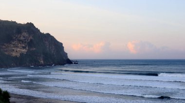 Tropikal plajın çarpıcı deniz manzarası kıyıya vuran dalgalar, dramatik sahil kayalıkları ve günbatımında pastel gökyüzü. Seyahat, doğa ve manzara için mükemmel..
