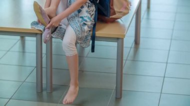 Bir kız bir bankta oturuyor ve terliklerini giyecek. İlk okul dersi yakında başlayacak..