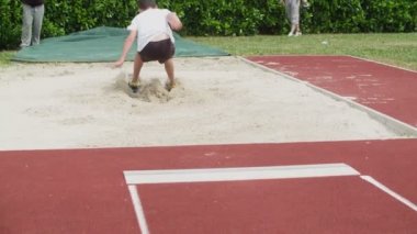 Bir öğrenci uzun bir atlayış yaptıktan sonra iki ayağına da iner. Başka bir öğrencinin peşinden atlayabilmesi için kum havuzundan hızla uzaklaşıyor..