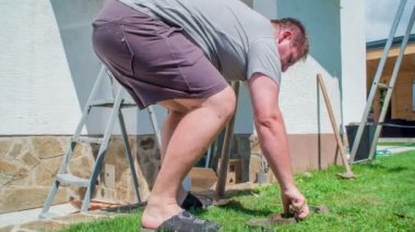 Genç adam çimlerdeki bakır parçalarını eğip topluyor ve onları bakır ürünleriyle dolu bir yığına koyuyor..