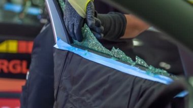 Bir tamirci kırık bir arabanın camını tamir etmeye başlamak üzere. O cam kırık parçaları tutar ve bir araba tamir dükkanında yere atar. Güvenlik eldiveni giyiyor..