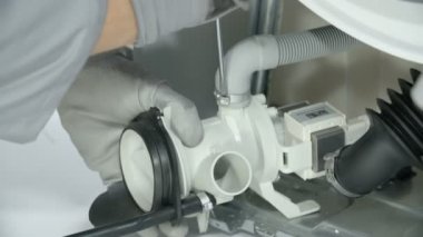 Tamirci bozuk çamaşır makinesini tamir ediyor. Şarjörü genişleterek pompanın hortumunu çıkartıyor..