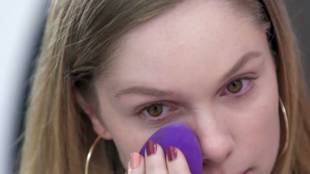 一个年轻的女人在她的脸上化妆 她将使用液体粉末 并使用软软的紫色海绵 — 图库视频影像
