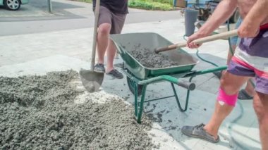 Yeşil metal bir el arabasıyla beton karışımı ve suyu çapayla karıştıran bir adam. Diğer iki adam dinleniyor ve aktiviteyi gözlemliyor..