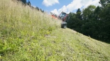 Bir traktör yavaş ve dikkatli bir şekilde yokuş aşağı sürüş ve tarım makineleri ile çim kesiyor. Gün güneşli ve sıcak.