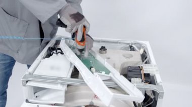 Bir tamirci kontrol panelindeki vidaları çıkarmak için matkap kullanıyor. Çamaşır ve kurutma makinesini tamir ediyor ve güvenlik kıyafetleri ve eldivenler giyiyor..