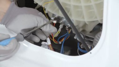 Bir tamirci çamaşır makinesinin arka stoktan kablolarını çekiyor. Tornavida ve güvenlik eldiveni kullanıyor..