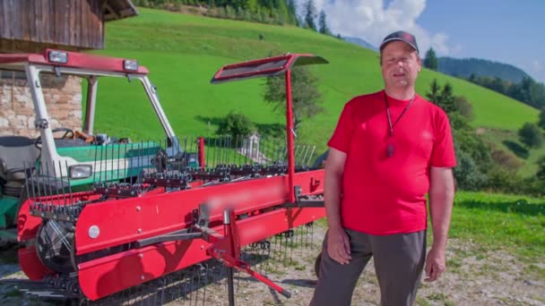 농부가 카메라에 무언가를 말하고 배경에서 건초를 돌리는 기계가 있습니다 화창하고 — 비디오