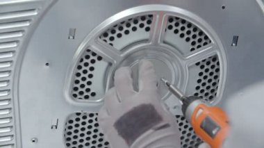 Bir adam vidaları tekrar takıyor. Matkap kullanıyor. Yıkama ve kurutma makinesinin tamir edilmesi gerekir.