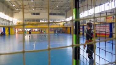 Bir kaleci kalenin önünde duruyor, böylece karşı takım topu kalesine atmıyor. Çocuklar bir okul spor salonunda futbol oynuyorlar..