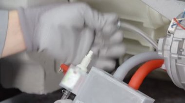 Bir adam çamaşır makinesinde birkaç tüpün fişini çekiyor. Tamir ediyor. Güvenlik eldiveni giyiyor..