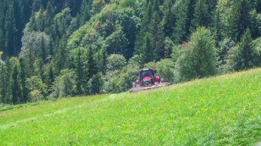 Bir çiftçi traktörde oturuyor ve çim kesme makineleriyle ot kesiyor. Gün güneşli ve sıcak.