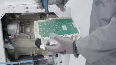 Bir adam çamaşır ve kurutma makinesitamir ediyor. Güvenlik kıyafetleri ve eldivenler giyiyor..