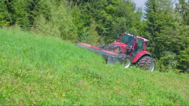 Bir traktör tepede sürüyor ve çiftçi çim kesme makineleri ile çim kesiyor. Gün güneşli ve güzel.
