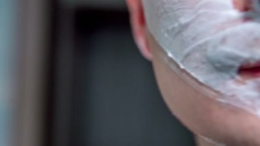 Genç bir adam banyoda yüzünü tıraş ediyor. Yüzünde bir sürü köpük var..