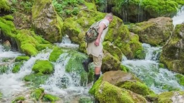 Bir adam yosunla kaplı büyük kayaların üzerinde yürüyor. Sonra orada durur ve biri onun fotoğrafını çeker..