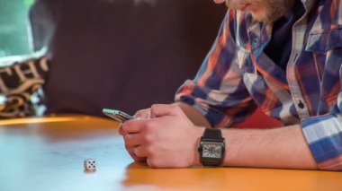 Genç bir adam masada oturuyor ve telefonundan bir şeyi kontrol ediyor. Masanın üzerinde beyaz bir zar da var..