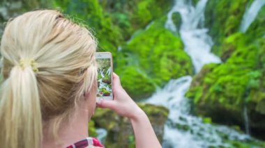 Sarı saçlı genç bir kadın şelalenin fotoğrafını çekiyor. Doğa güzel ve yeşil.