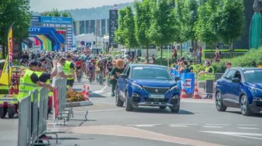 Turnuvaya yeni başlayan iki büyük mavi araba bisiklet yarışçılarının önünde sürüyor..