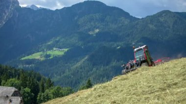 Bir traktör dik bir tepede saman hazırlıyor. Arka planda güzel dağlar var ve manzara harika..