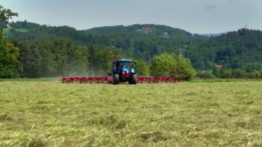 Büyük mavi bir traktör büyük bir tarladan geçiyor ve saman hazırlıyor. Hava güneşli. Arka planda tepeler var.