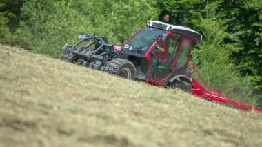 Kırmızı bir traktör yokuş yukarı gidiyor ve tarım makinelerinin yardımıyla samanları yığınlara ayırıyor..
