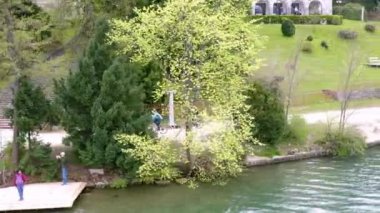Nehrin yanında güzel bir park görebiliyoruz ve insanlar etrafta yürüyüp doğanın tadını çıkarıyorlar. Arkadaki tepede büyük bir bina var..