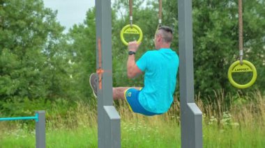 Bir sporcu dışarıda güç egzersizleri yapıyor. Yüzüklerle kendini kaldırıyor..